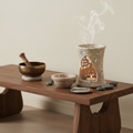 Brûleur d'huile essentielle en pierre savon avec soucoupe métallique, posé sur un meuble zen à côté d'un bol tibétain